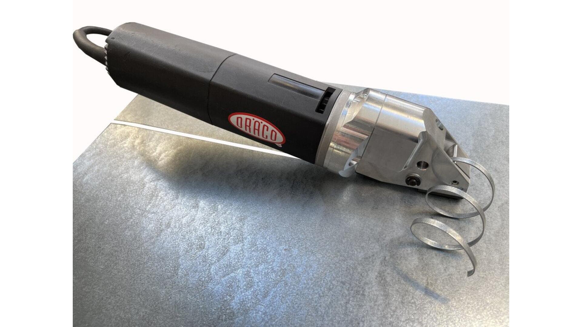 An electric metal sheet nibbler tool is positioned on a metallic surface, cutting a thin strip and creating curled metal shavings. The tool is labeled DRACO.