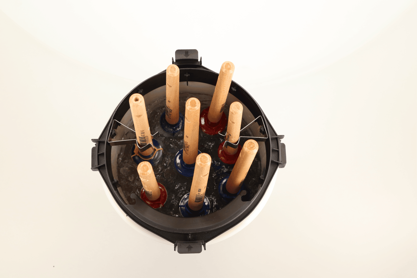 Top-down view of seven wooden sticks standing vertically in a circular black container, each inserted into a colored holder submerged in water, likely for cleaning or soaking purposes.