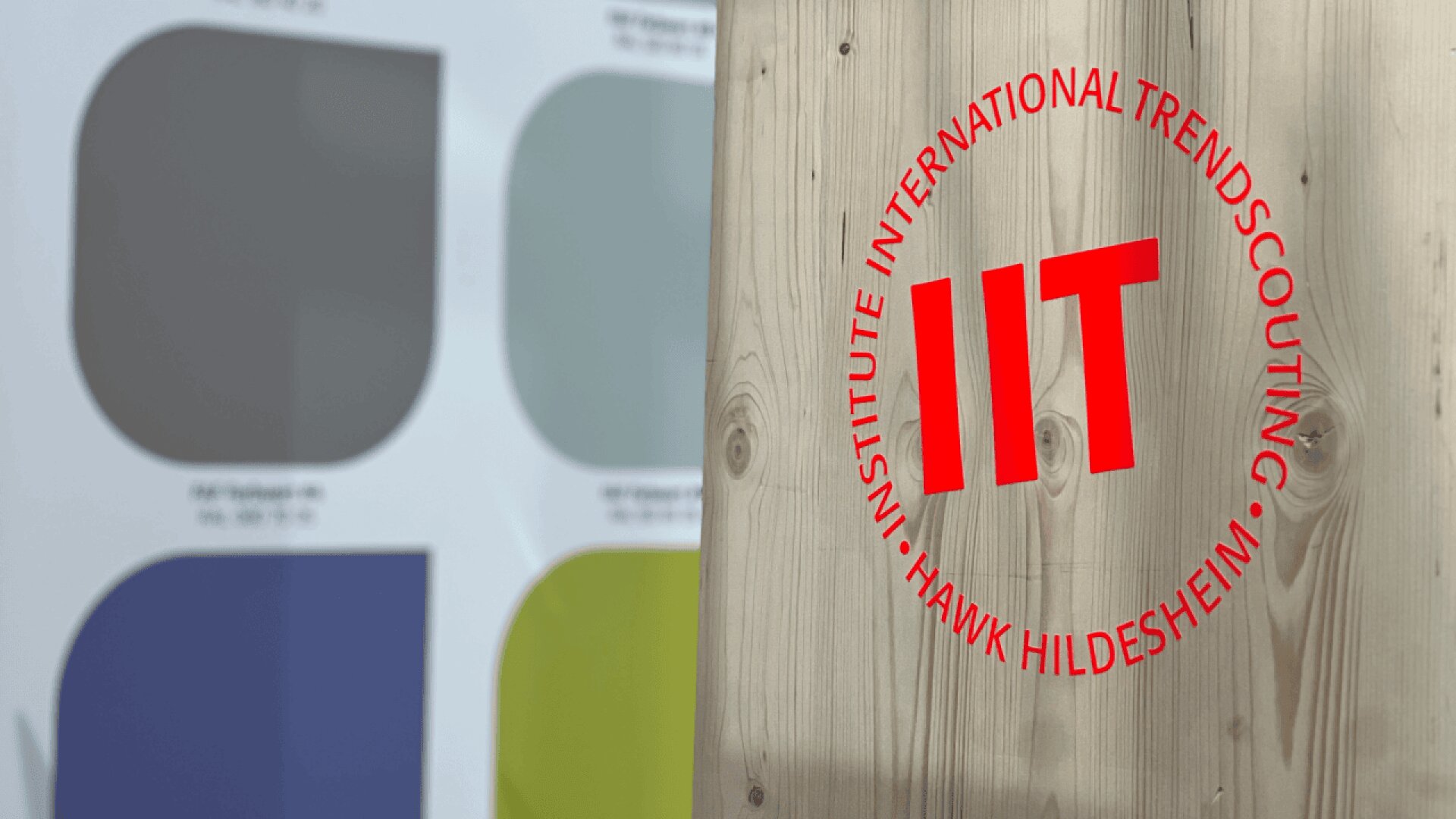 A wooden panel with a red IIT logo and text Institute International Trendscouting HAWK Hildesheim stands in front of a blurred background featuring large color swatches in gray, blue, and white.