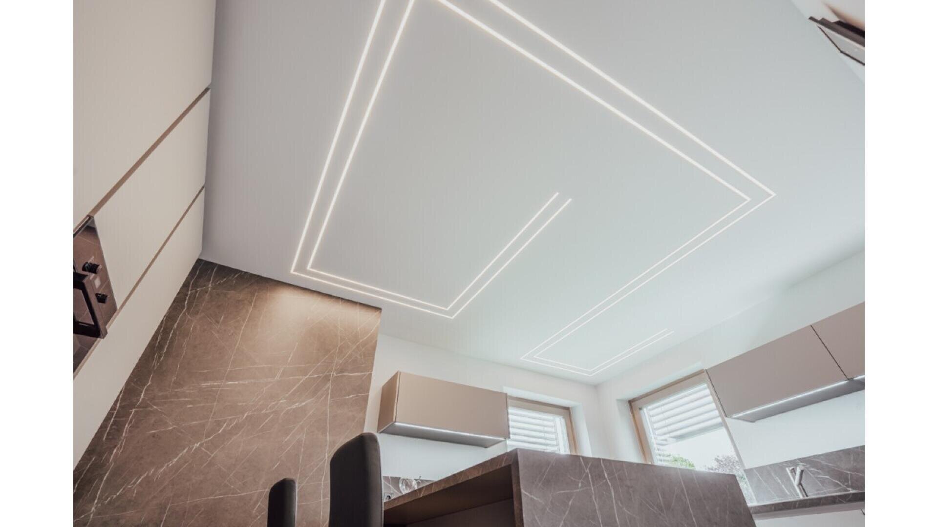 Modern kitchen with a marble-patterned wall, minimalistic white cabinets, and a unique ceiling design featuring geometric LED light strips. Large windows provide natural light, creating a bright and contemporary atmosphere.