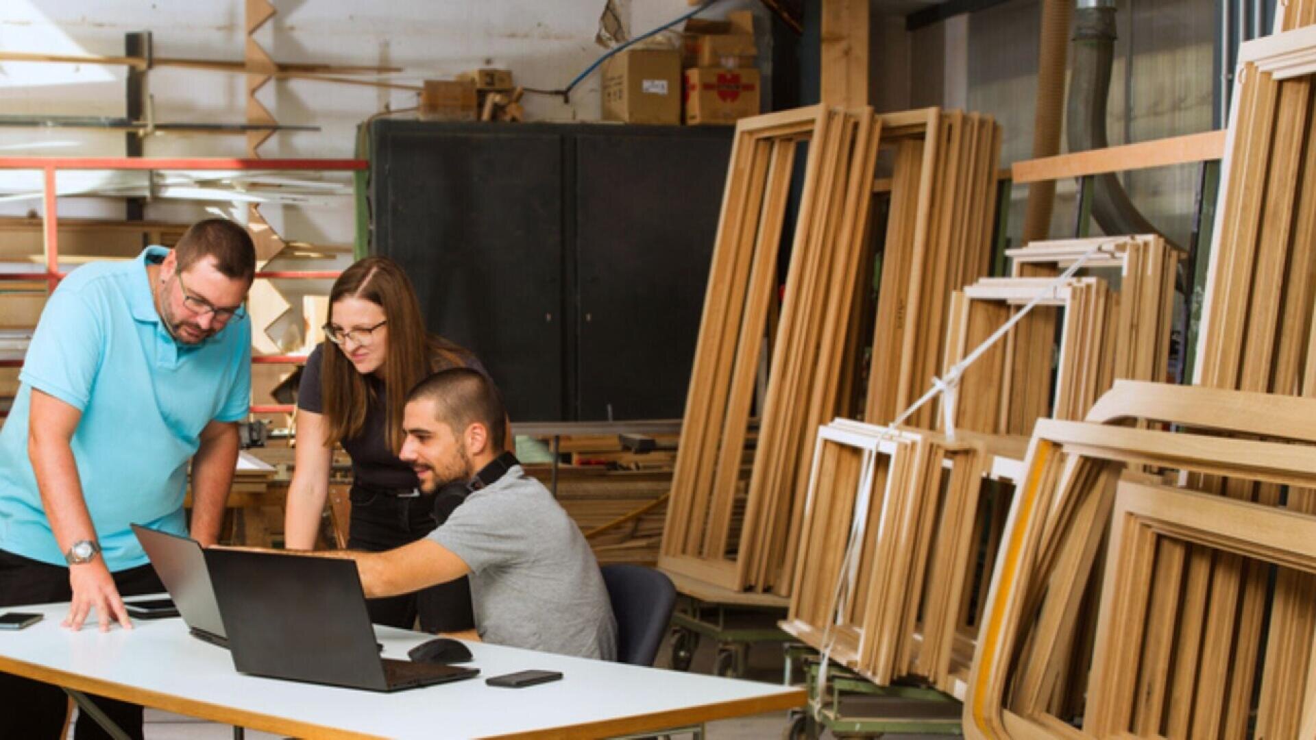 Three people in a woodworking shop look at laptops on a table, discussing something. Wooden frames are stacked nearby, and tools and materials are visible in the background.