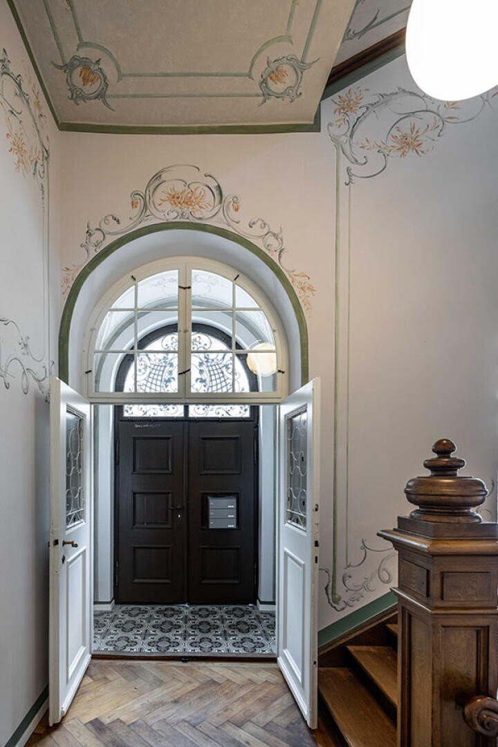 Eleganter Eingangsbereich mit Doppeltüren, die sich öffnen und den Blick auf eine dunkle Haupttür aus Holz, ein Bogenfenster darüber, kunstvolle Wand- und Deckengestaltungen, gemusterte Bodenfliesen und ein hölzernes Treppengeländer auf der rechten Seite freigeben.