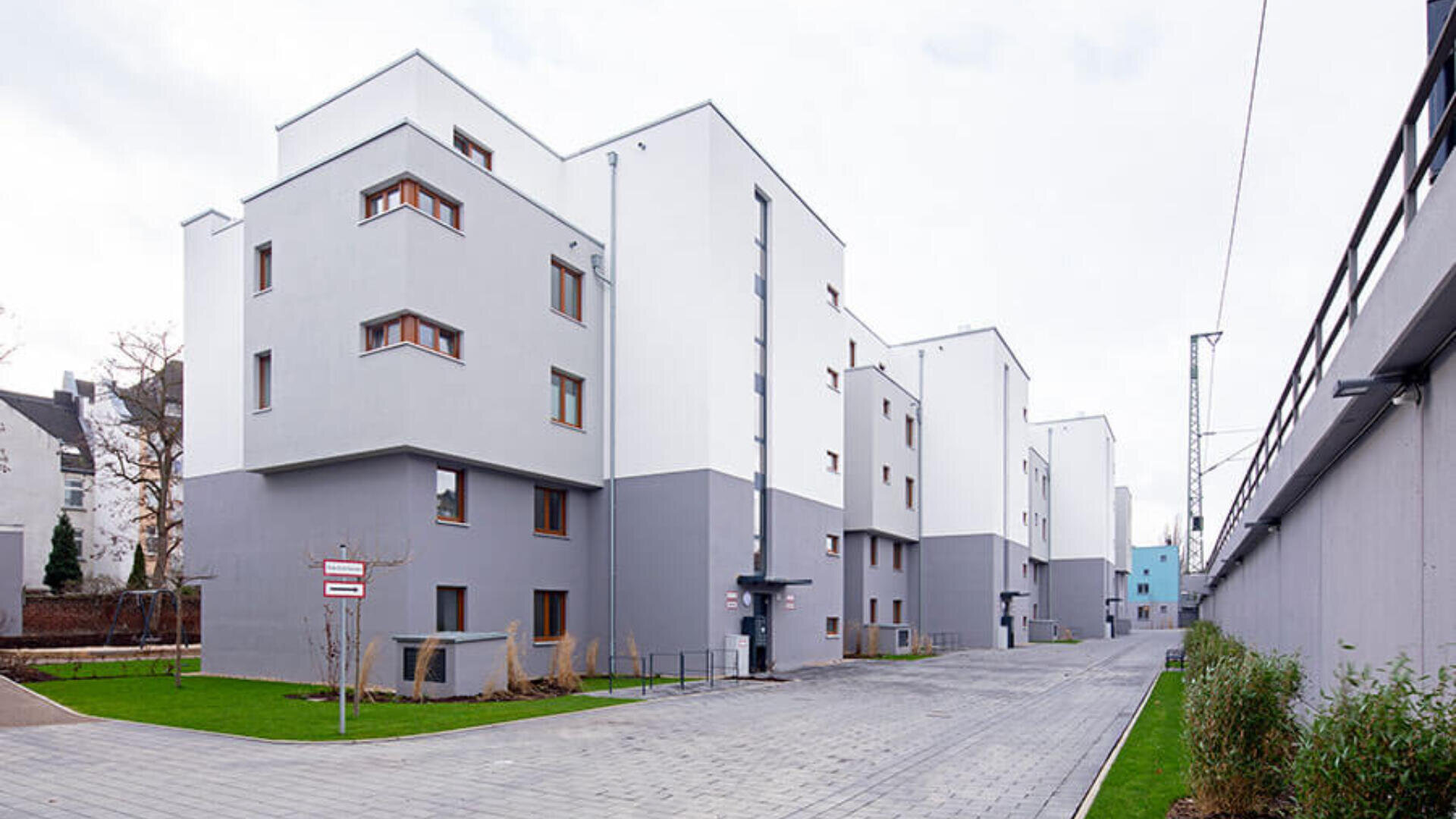 Eine Reihe moderner, weiß-grauer Wohnhäuser mit Flachdächern und hölzernen Fensterrahmen säumen einen sauberen, gepflasterten Gehweg, der von grünem Gras und kleinen Pflanzen gesäumt ist.