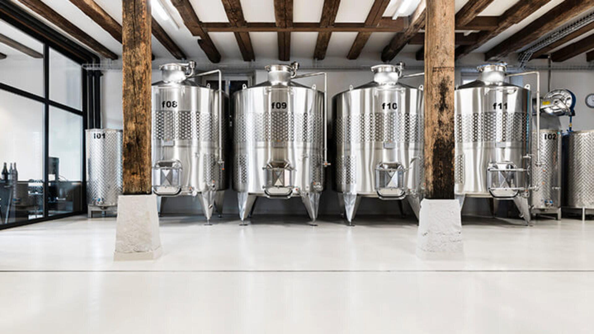 Vier große Gärtanks aus Edelstahl mit nummerierten Etiketten stehen in einer sauberen, modernen Industriehalle mit freiliegenden Holzbalken und gläsernen Bürowänden.