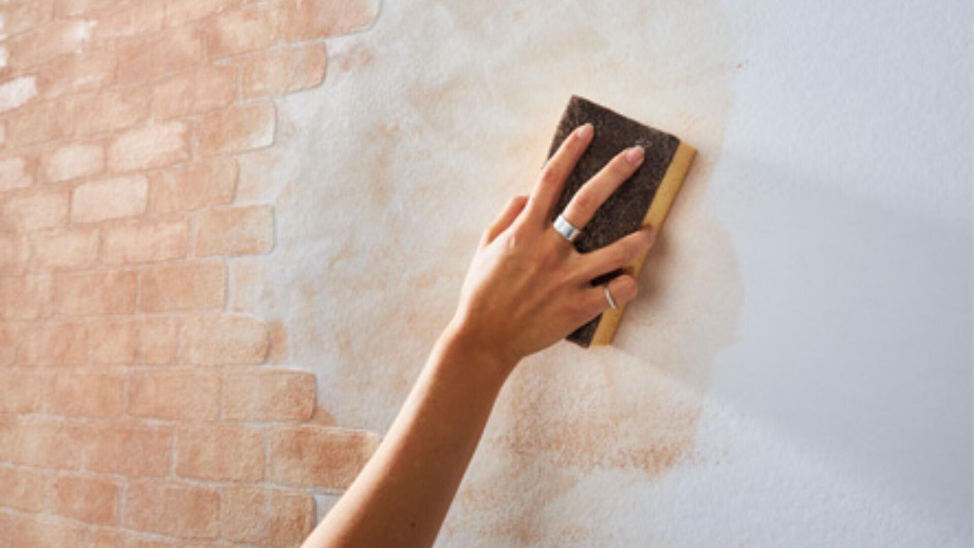 Eine Hand mit einem Ring reinigt oder überstreicht mit einem Schwamm eine mit Ziegelmuster gestaltete Wand und gibt dabei einen schlichten weißen Bereich frei.