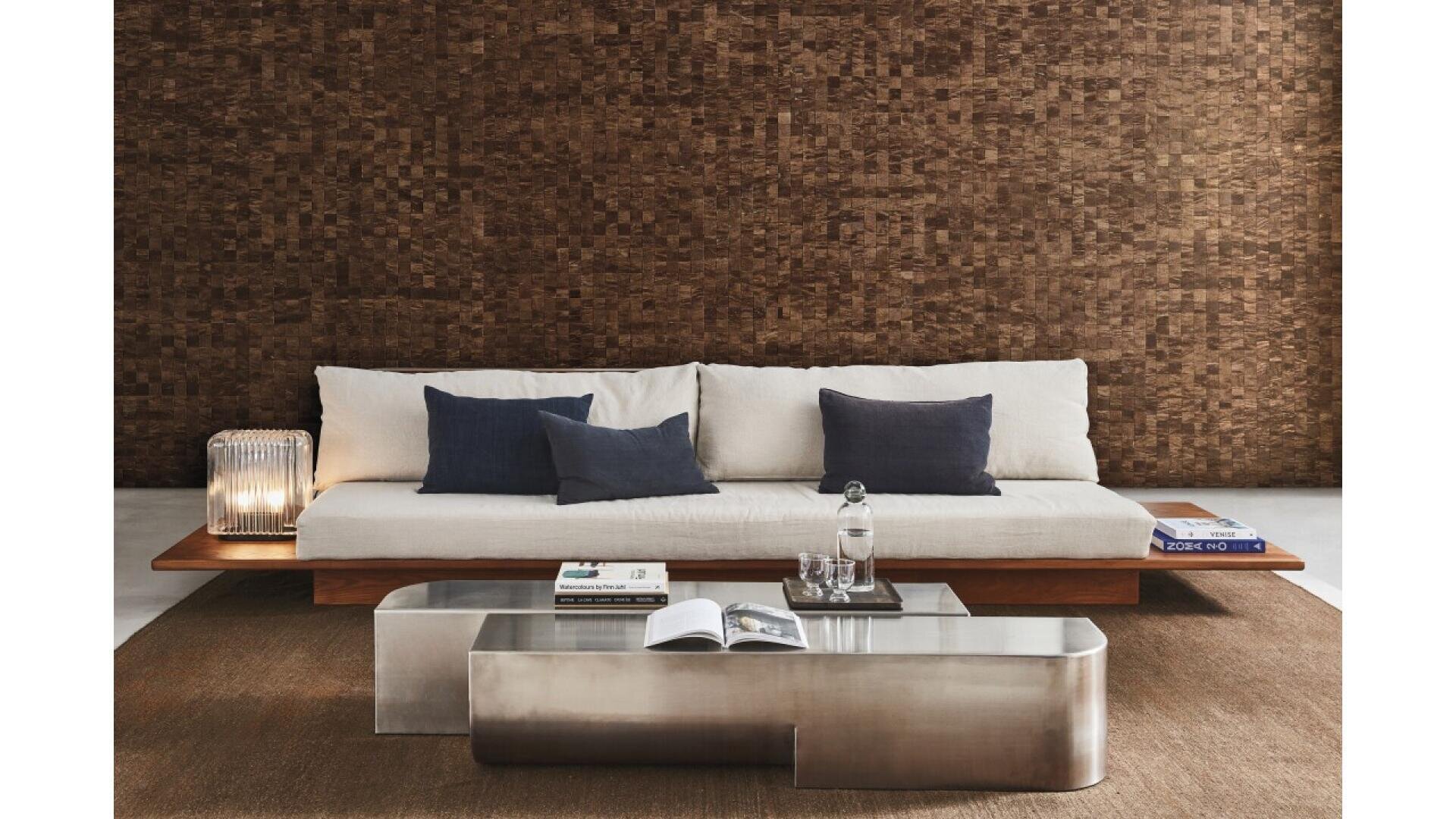 A modern living room with a low wooden sofa featuring white cushions and navy pillows, a clear glass lamp, two metallic coffee tables with books and a water carafe, and a brown textured wall backdrop.