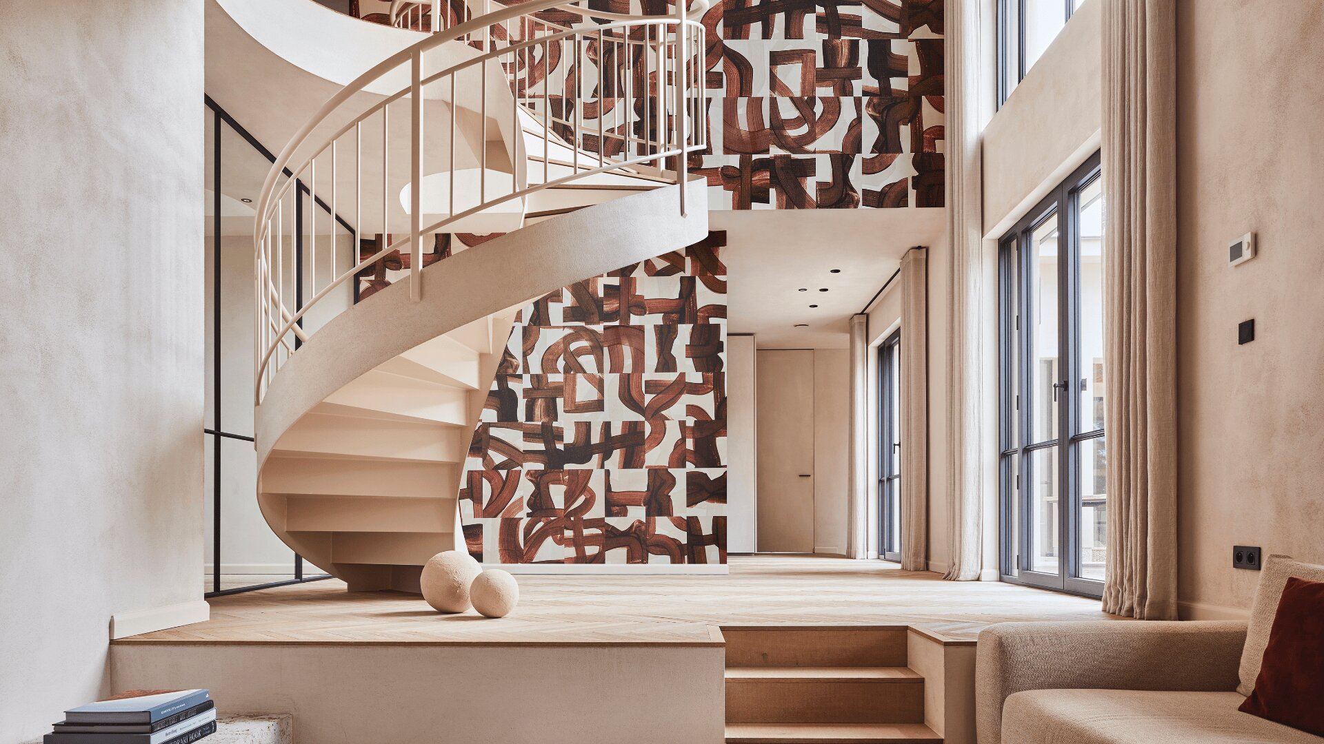 Ein modernes Interieur mit einer weißen Wendeltreppe, einer abstrakt gemusterten Akzentwand, großen Fenstern, neutral getönten Wänden und Möbeln sowie zwei dekorativen Kugeln auf einem hellen Holzboden.