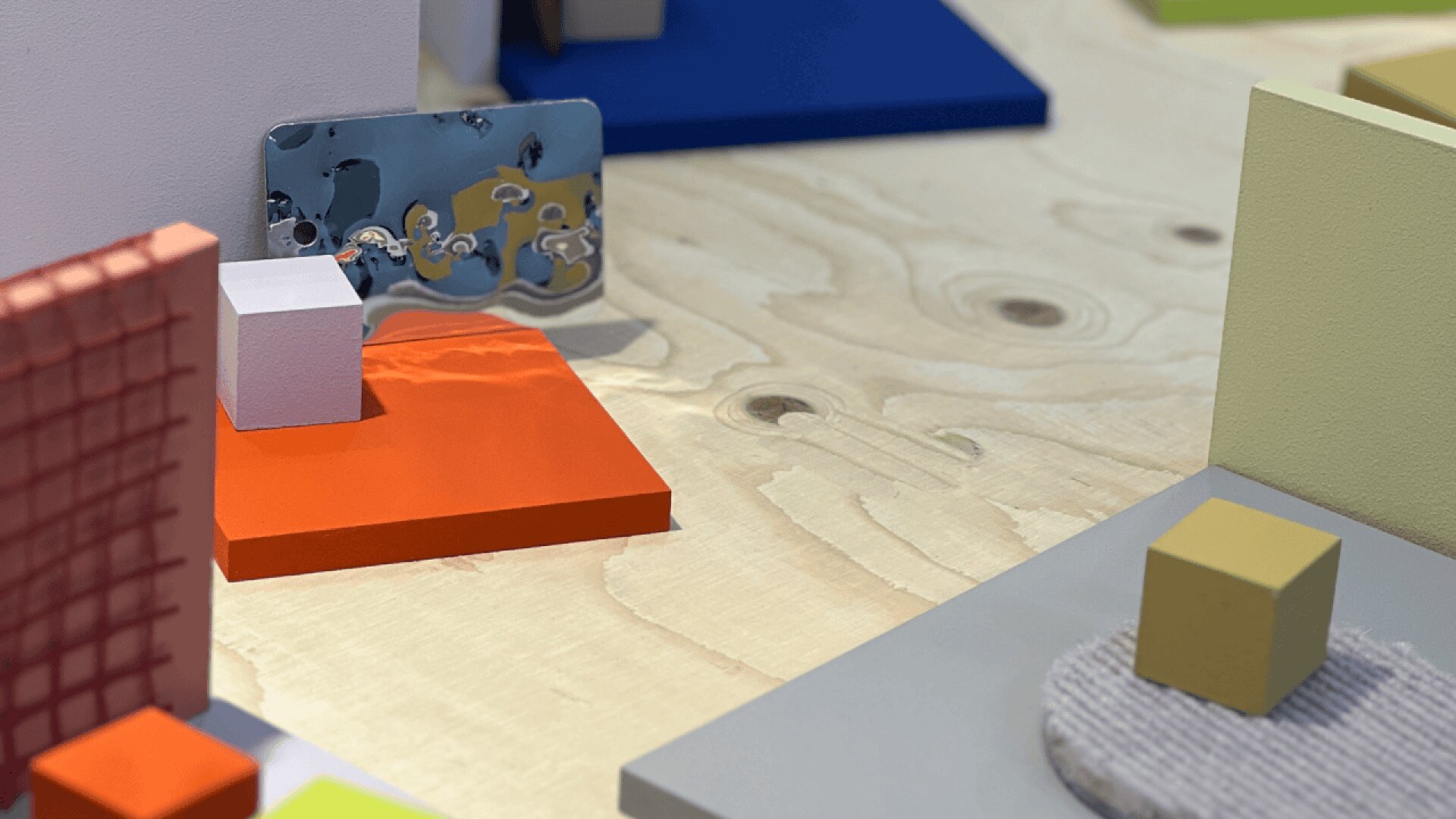 Colorful geometric shapes, including cubes and rectangles, are arranged on a light wooden surface. A reflective, metallic object with holes is positioned near the center, surrounded by orange, blue, and gray platforms.