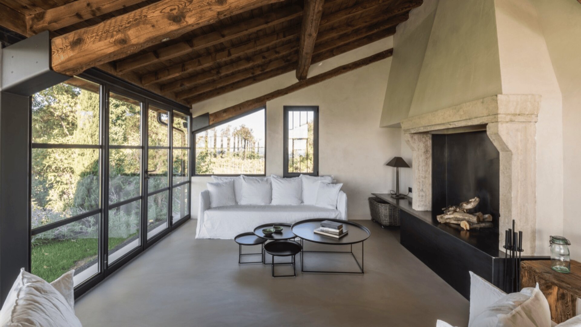 Ein gemütliches Wohnzimmer mit schräger Holzdecke, großen Fenstern, einem weißen Sofa, verschachtelten runden Couchtischen und einem Kamin mit Holzstapeln. Natürliches Licht durchflutet den Raum und unterstreicht die moderne rustikale Einrichtung.