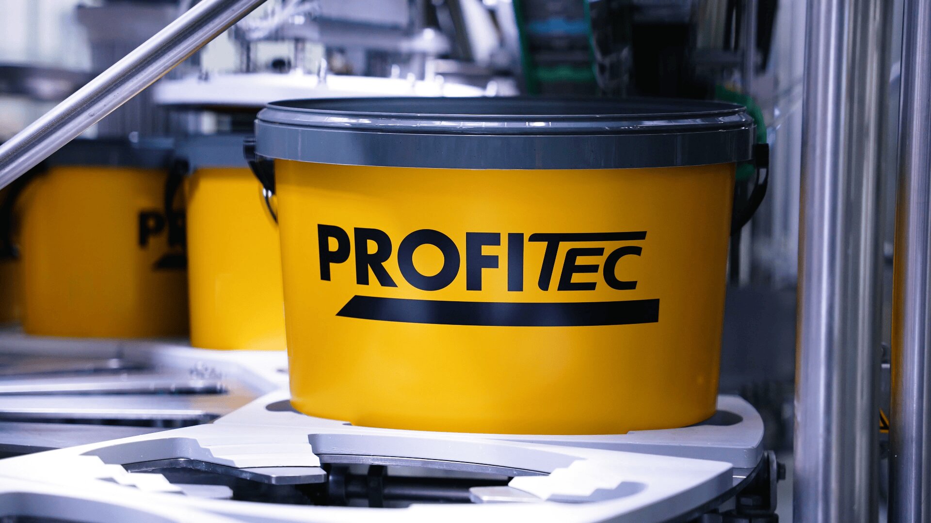 A yellow plastic bucket with a gray lid and the word PROFITEC printed in bold black letters is placed on industrial equipment in a manufacturing setting.