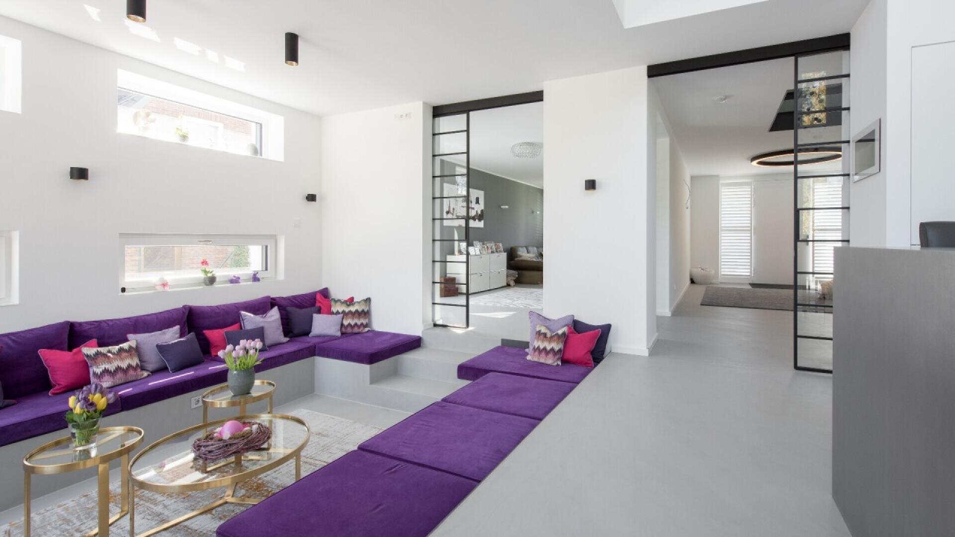 Modern living room with a sunken seating area featuring purple cushions, colorful accent pillows, and glass coffee tables with flowers, surrounded by white walls and large windows allowing natural light in.