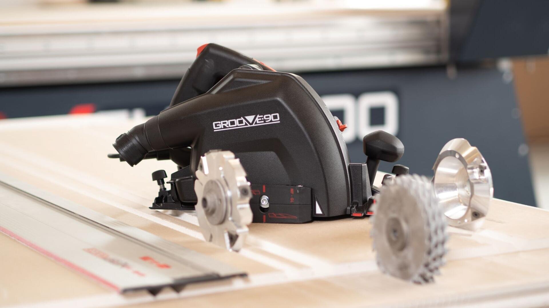A black GROOVE 90 power tool is placed on a work surface with two circular saw blades nearby. The background shows part of another machine, slightly out of focus.