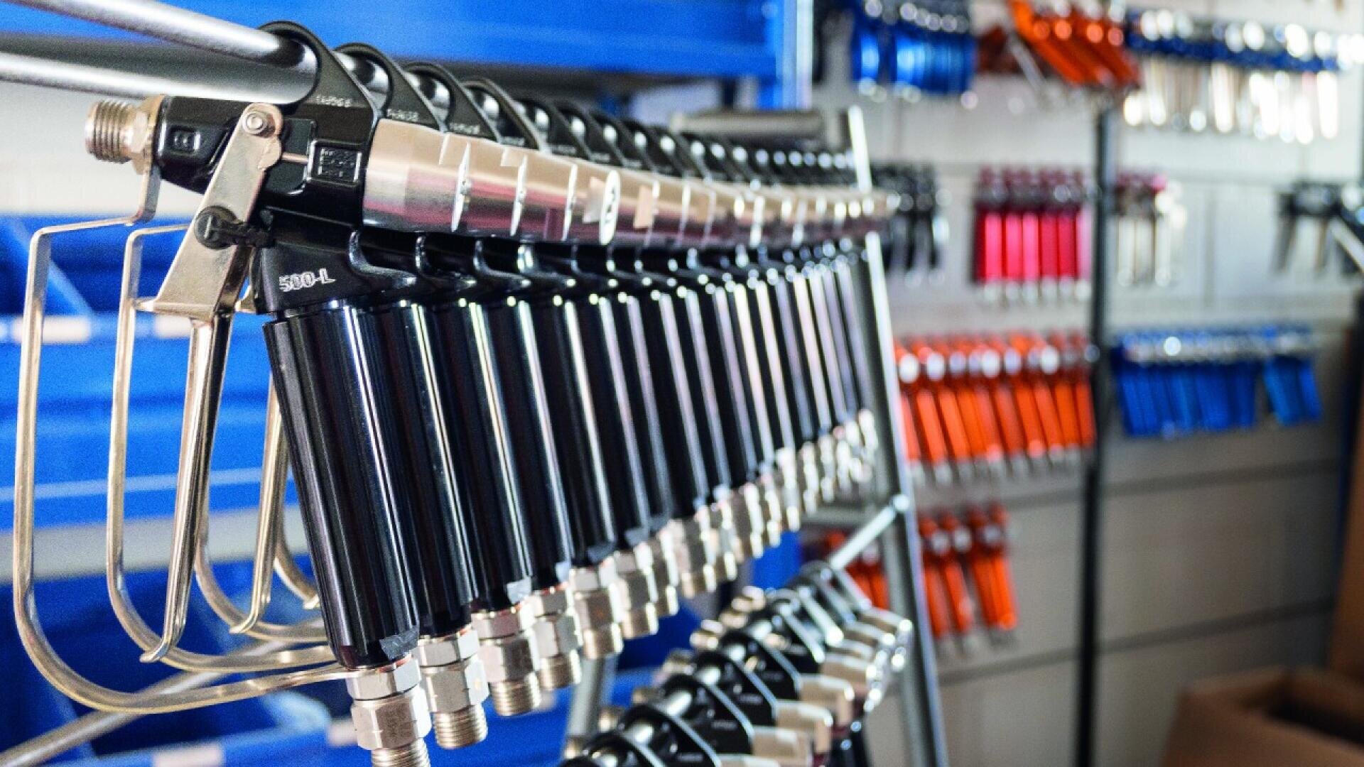 A row of black spray guns is hanging on a metal rack in the foreground, with more spray guns in various colors organized on shelves in the background. The setting appears to be a workshop or hardware store.