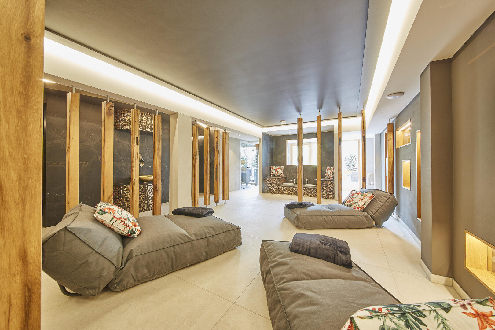 Eine moderne Entspannungslounge mit grau gepolsterten Chaiselongues, geblümten Kissen, hölzernen Raumteilern und sanfter, indirekter Beleuchtung. Große Fenster lassen natürliches Licht herein und schaffen eine ruhige und einladende Atmosphäre.