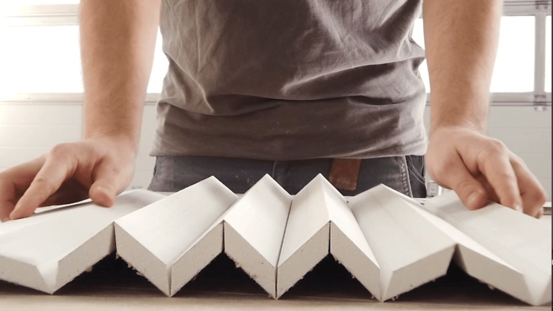 Eine Person in einem grauen Hemd ordnet mehrere weiße, zickzackförmige Tafeln oder Kacheln auf einer ebenen Fläche an, die aus einem niedrigen Winkel betrachtet wird.