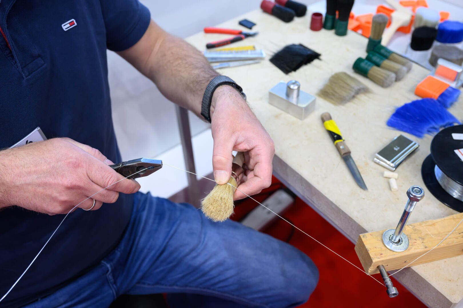 Eine Person befestigt mit einer Zange Draht um ein Borstenbündel und baut an einer Werkbank, die mit Werkzeugen, Bürsten und Materialien bedeckt ist, eine Bürste zusammen.
