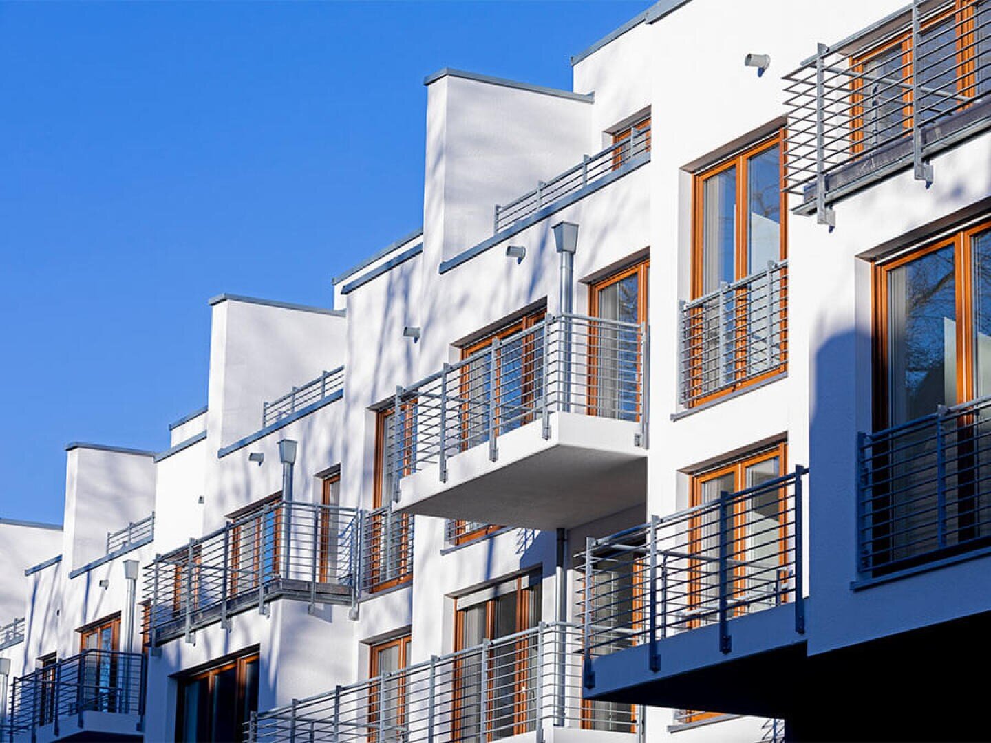 Modernes weißes Mehrfamilienhaus mit Holzfenstern und Metallgeländern an den Balkonen vor einem strahlend blauen Himmel. Die Architektur zeichnet sich durch klare Linien und geometrische Winkel aus.