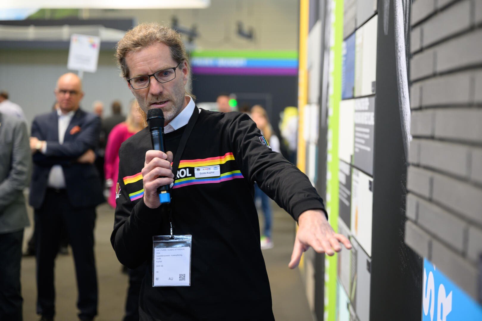 Ein Mann mit Brille und Mikrofon deutet bei einer Indoor-Veranstaltung auf eine Anzeigetafel. Im Hintergrund beobachten Menschen in Business-Kleidung die Veranstaltung.
