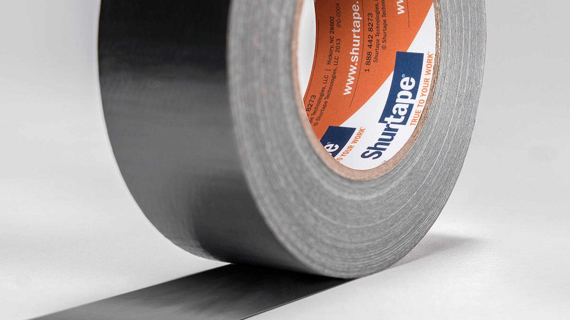 Eine Rolle schwarzes Shurtape-Klebeband wird auf einer weißen Oberfläche abgerollt, wobei das Band teilweise festklebt und das Markenetikett sichtbar ist.