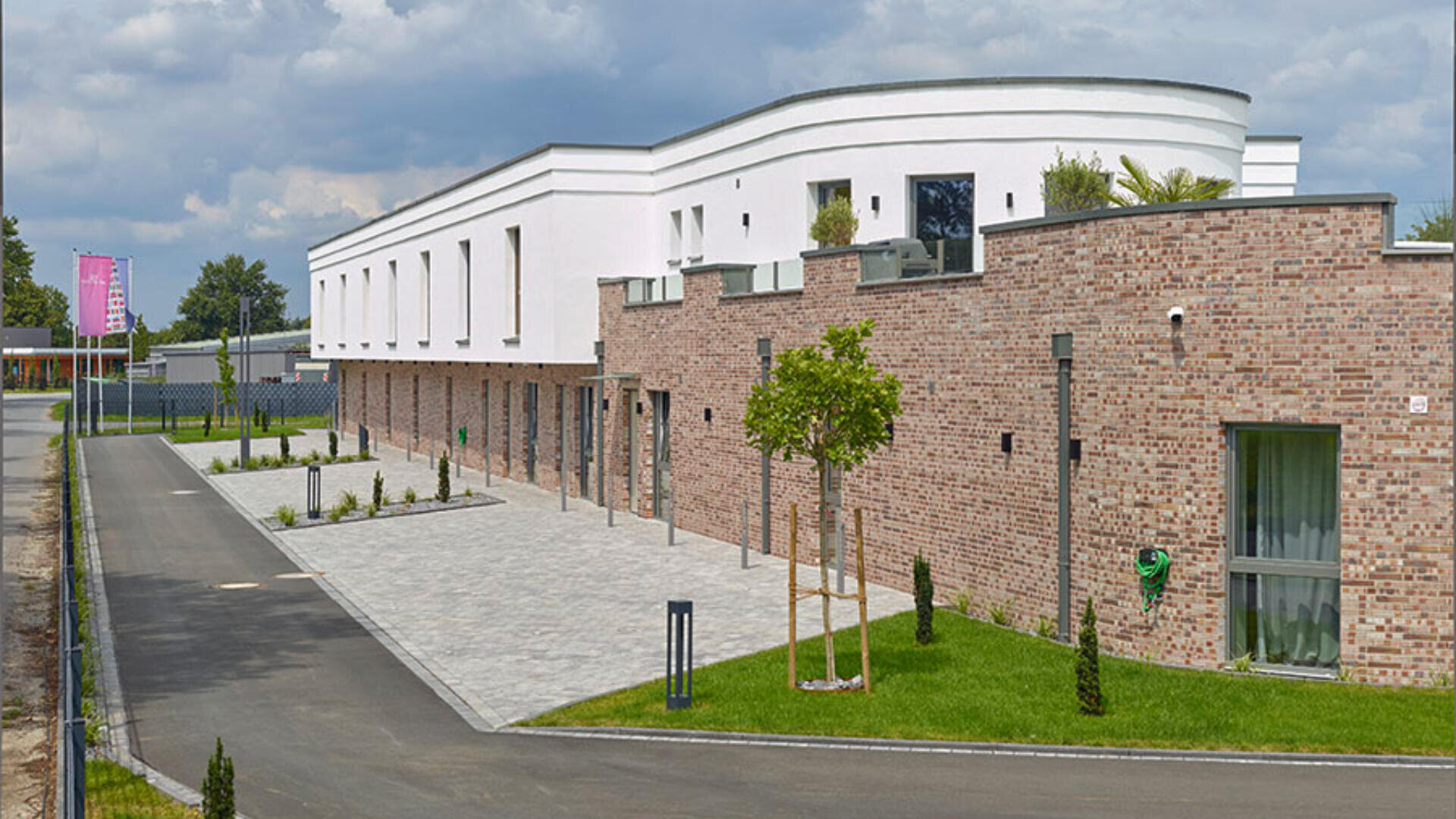 Ein modernes Gebäude mit weißen Wänden und einem geschwungenen Dach steht unter einem bewölkten Himmel vor einem Hintergrund aus braunen Backsteinmauern, gepflegtem Rasen, jungen Bäumen und einem Steinweg.