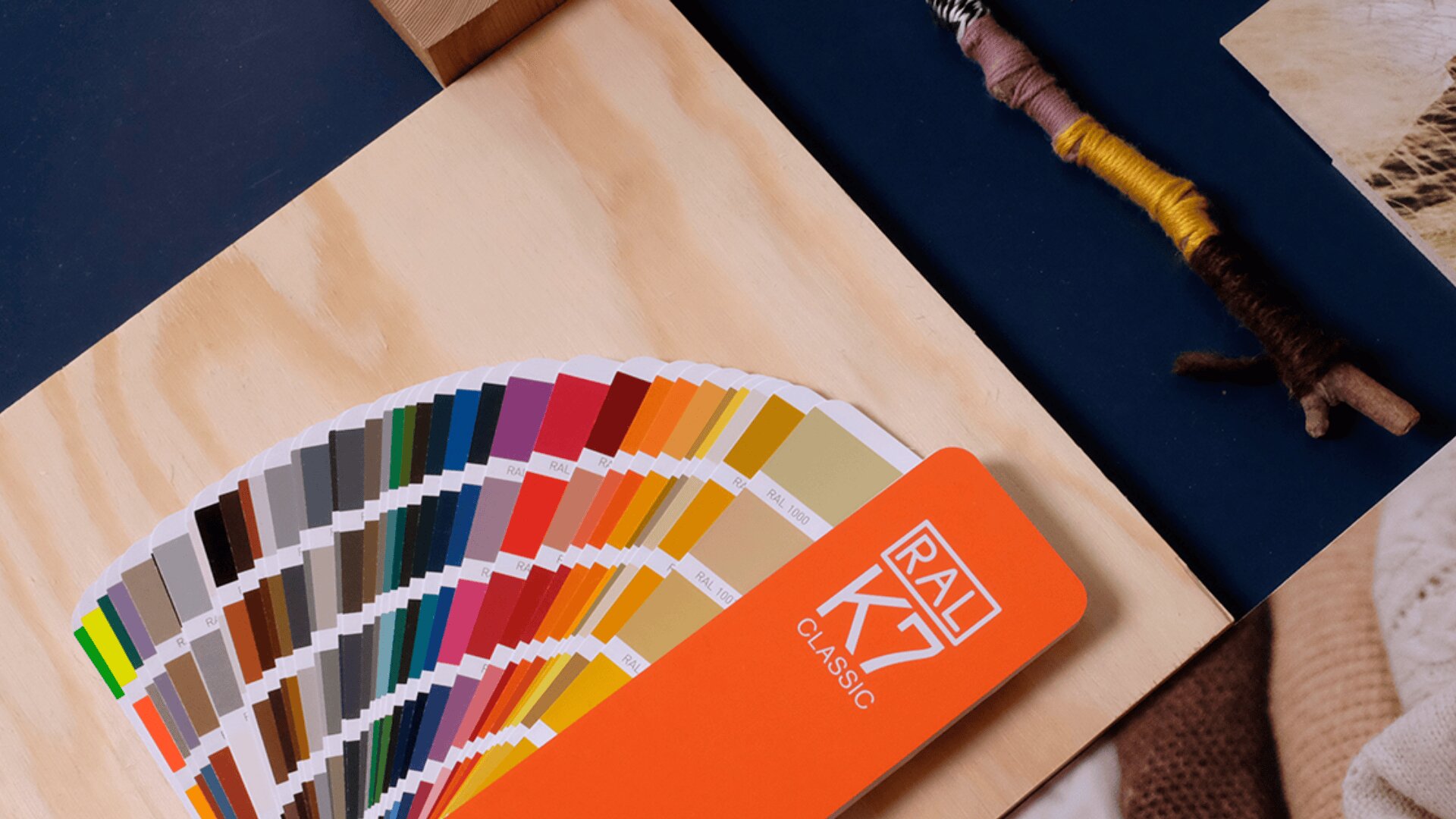 Auf einer hellen Holzfläche ist ein Farbfächer RAL K7 Classic ausgebreitet, der verschiedene Farbmuster zeigt. Daneben liegen mit Garn umwickelte Stäbchen und andere Kreativmaterialien.