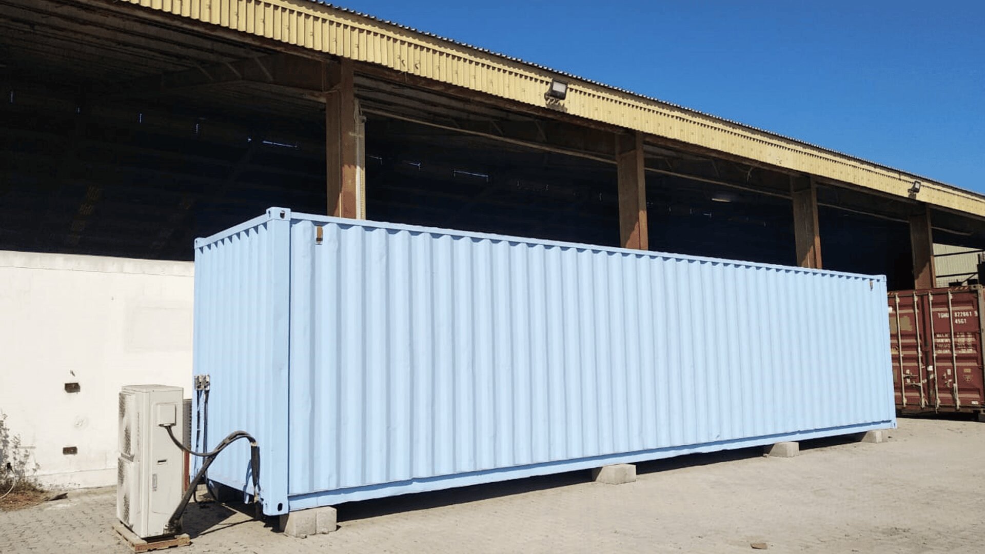 Ein großer hellblauer Schiffscontainer steht auf Betonblöcken vor einer Industriehalle mit gelbem Dach, neben einer weißen Wand und einer Klimaanlage. Der Himmel ist klar und blau.