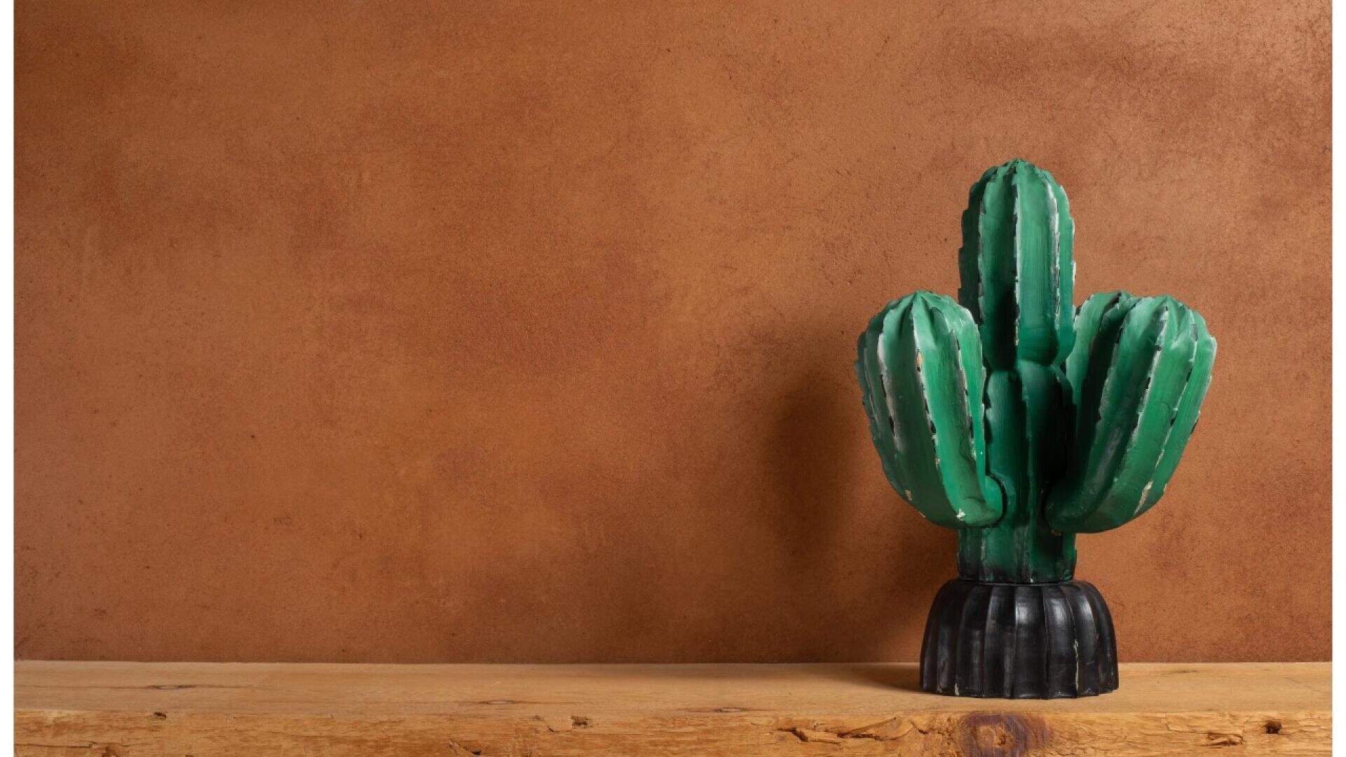 A green cactus sculpture with three arms stands on a wooden surface against a plain brown textured wall. The cactus has a black base and is centered to the right of the image.