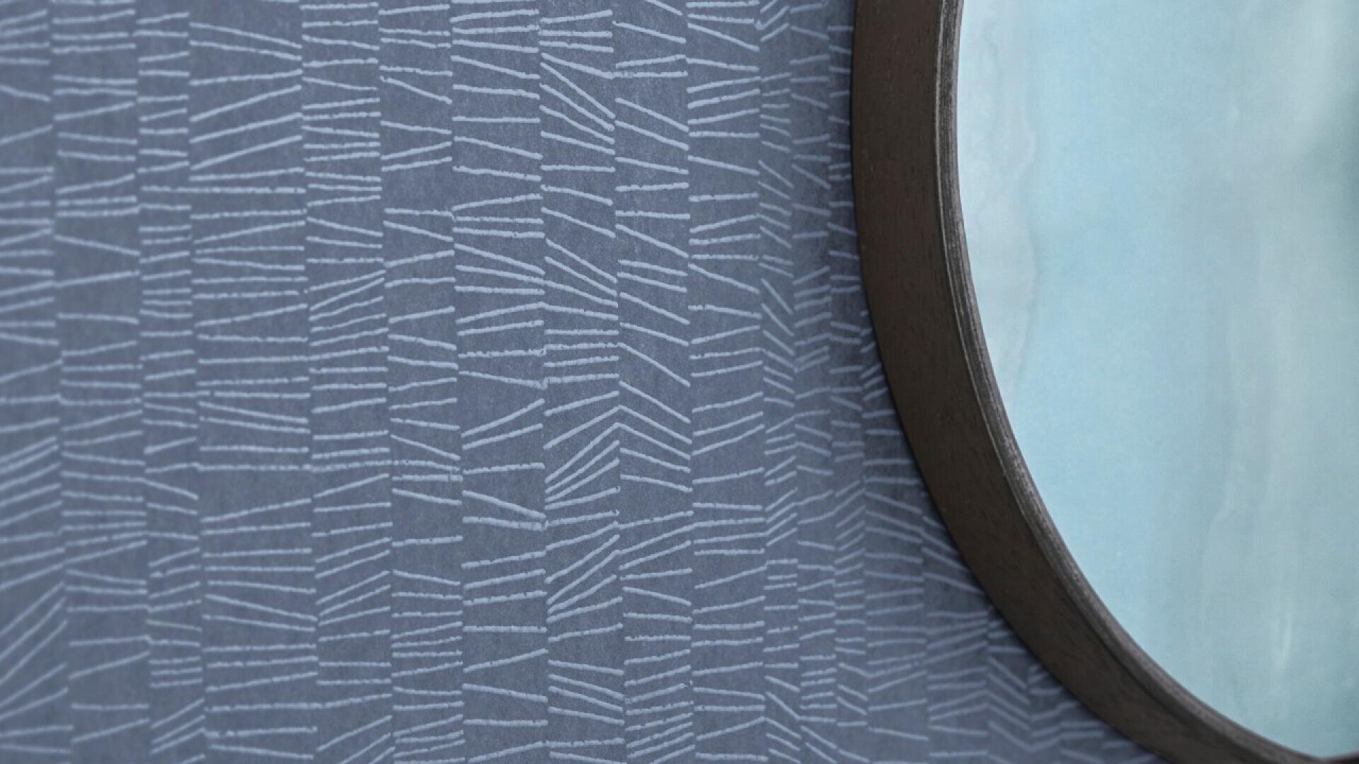 A close-up of a patterned gray-blue wall with white geometric lines and the edge of a round mirror with a dark frame, reflecting a light blue surface.