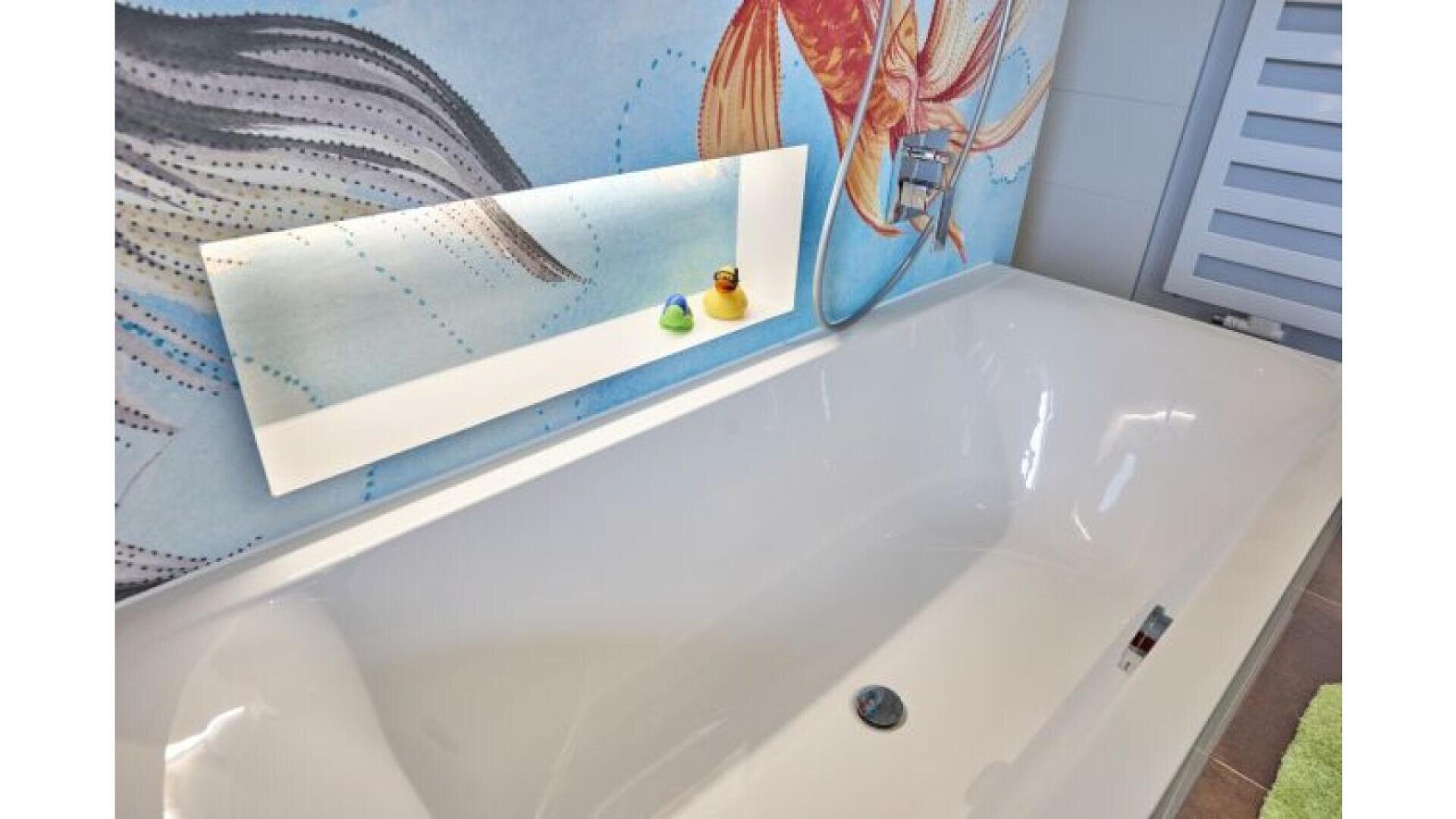 A white bathtub with a built-in shelf above it holding two rubber ducks, set against a colorful mural featuring a fish and abstract aquatic designs.