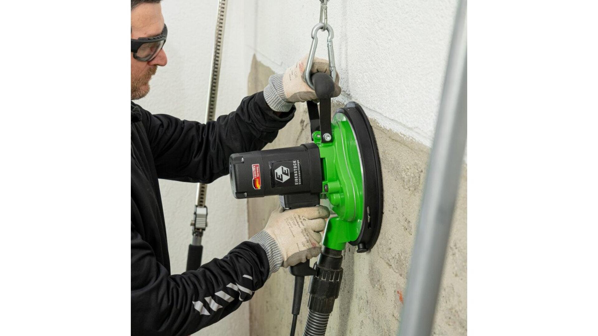 A person wearing gloves and safety glasses operates a green wall grinder or polisher, attached to a vacuum hose, smoothing the surface of a concrete wall.