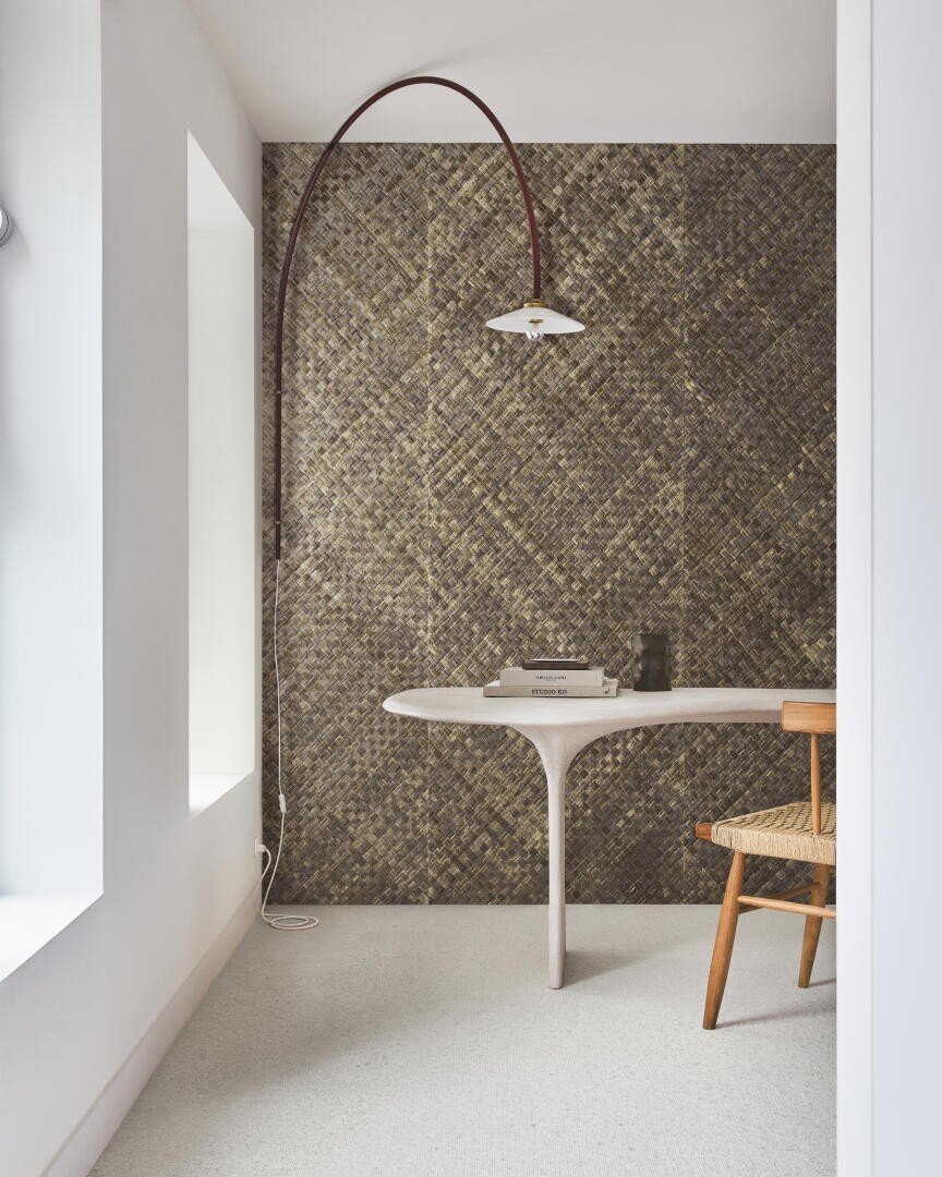 Ein moderner, minimalistischer Arbeitsplatz mit einem geschwungenen weißen Schreibtisch, einem Stuhl aus geflochtenem Holz, einer hohen, gewölbten Stehlampe und einer strukturierten Mosaik-Akzentwand in Gold- und Brauntönen. Licht strömt durch ein großes Fenster herein.