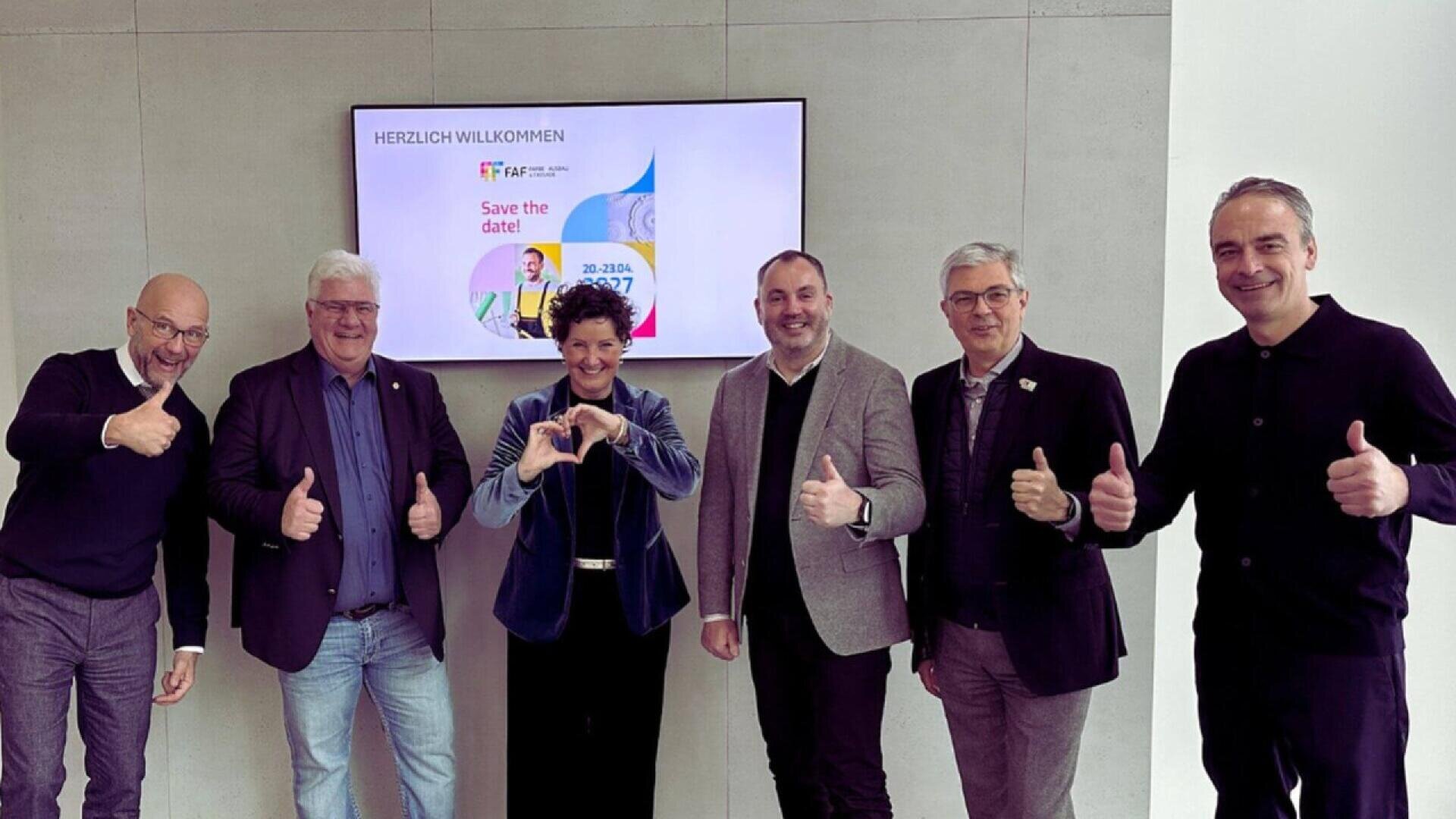 Six people stand in front of a screen, smiling and posing with thumbs up and a heart gesture. The screen behind them displays a welcome message and a “Save the date!” announcement.