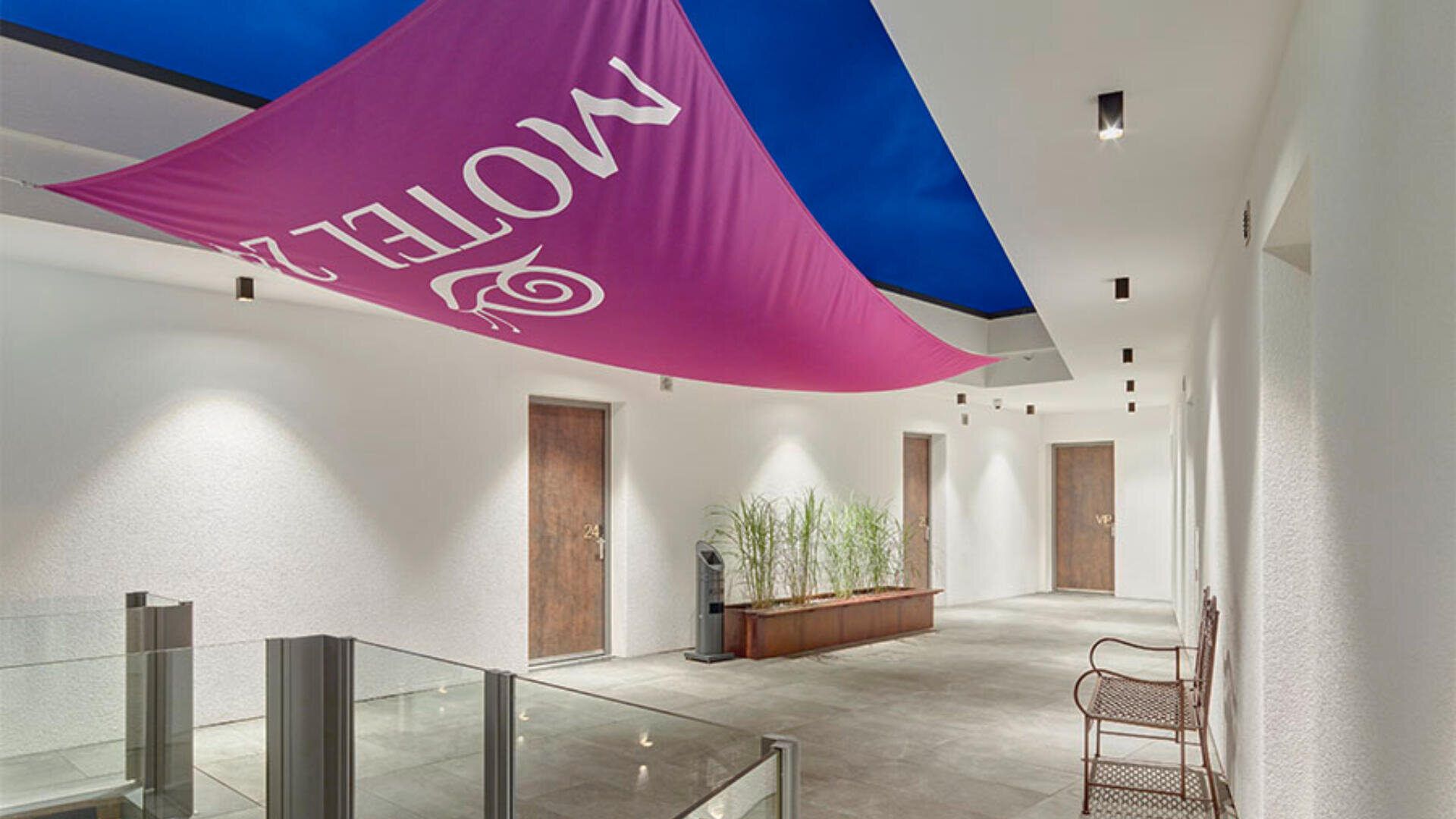 Ein moderner Hotelflur mit drei Holztüren, einem rosa Stoffbanner mit der Aufschrift „MOTEL“, das von der Decke hängt, Topfpflanzen, einer Metallbank und weißen Wänden unter einer blauen Decke.