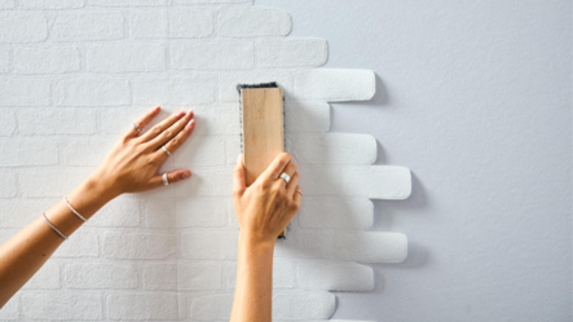 Eine Person streicht eine weiße Strukturtapete mit einem Holzwerkzeug glatt an eine Wand, wobei sie mit einer Hand die Tapete hält und mit der anderen Hand das Werkzeug dagegen drückt.