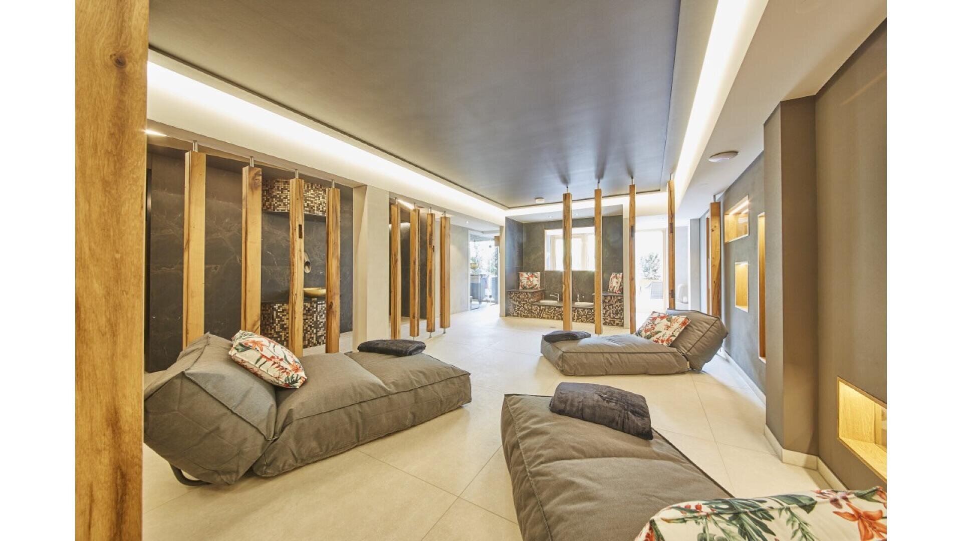 A modern relaxation lounge with cushioned gray lounge chairs, decorative wooden dividers, soft lighting, and floral pillows, creating a cozy and serene atmosphere. Large windows provide natural light in the background.