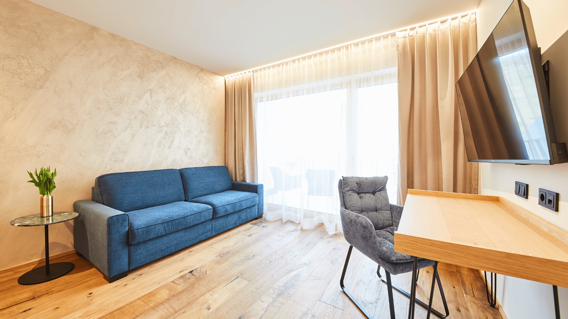 Modernes Wohnzimmer mit blauem Sofa, rundem Glastisch mit grünen Blumen, Holzschreibtisch und grauem Stuhl, großem Fenster mit beigen Vorhängen und Wandfernseher. Der Raum verfügt über helle Holzböden.