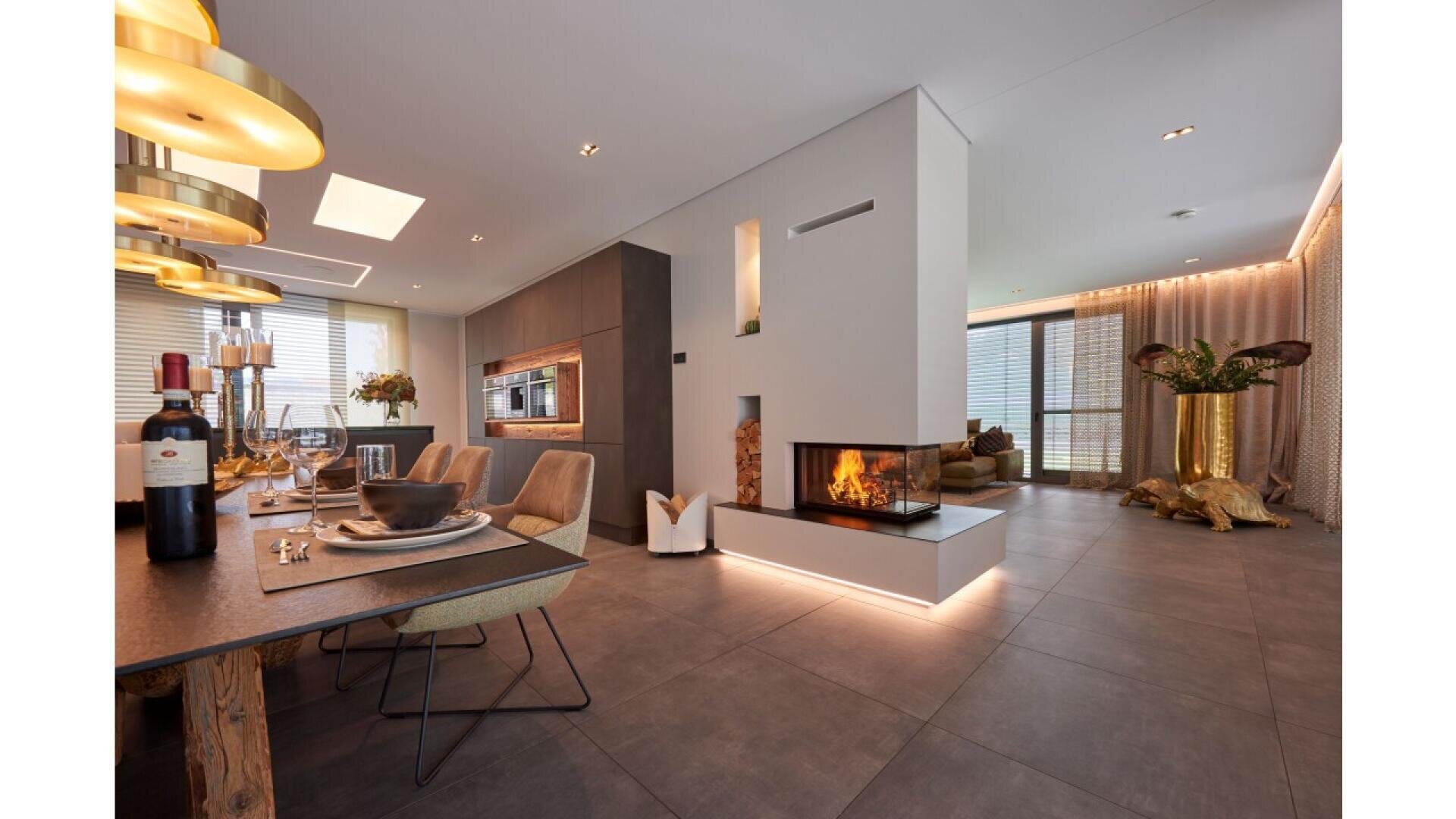 Modern open-plan dining and living area with a central double-sided fireplace, elegant lighting, set dining table, large windows, and decorative sculptures, including a gold animal figure and large vases with plants.