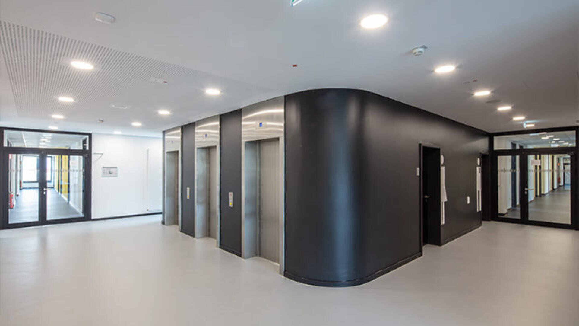 Ein moderner, gut beleuchteter Flur mit drei Aufzügen aus Edelstahl in einer geschwungenen schwarzen Wand, umgeben von weißen Böden und Decken sowie Glastüren, die zu anderen Fluren führen.