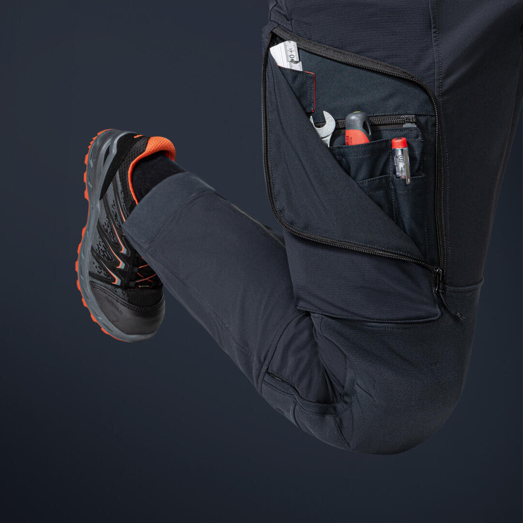 Eine Person in dunklen Hosen und schwarzen Wanderschuhen mit orangefarbenen Akzenten beugt ihr Knie und gibt den Blick auf eine Reißverschlusstasche am Oberschenkel frei, in der sich ein Stift, eine kleine Taschenlampe und andere Werkzeuge befinden.