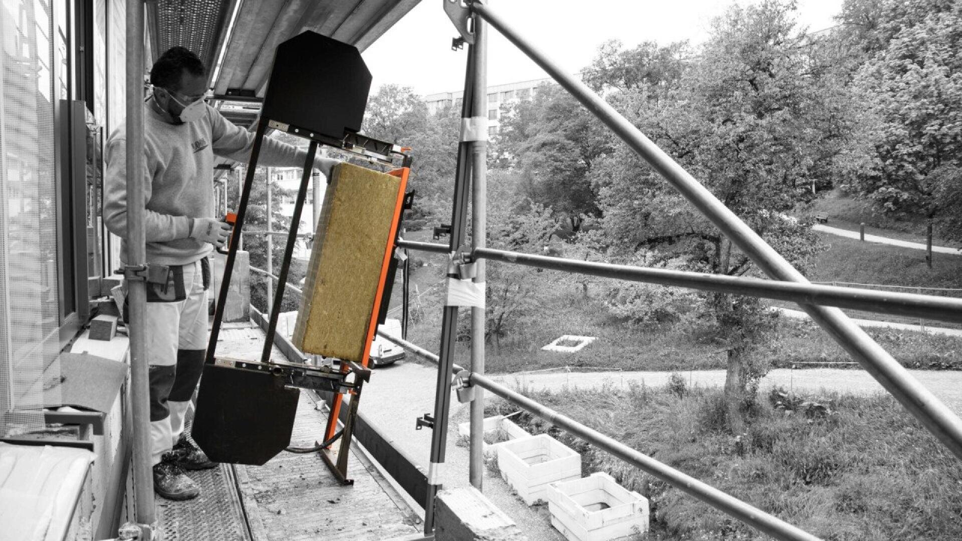 A worker on scaffolding adjusts a black and orange frame with a board, beside a building under construction. The background shows trees and a grassy area. The image is mostly black and white except for the frames color.