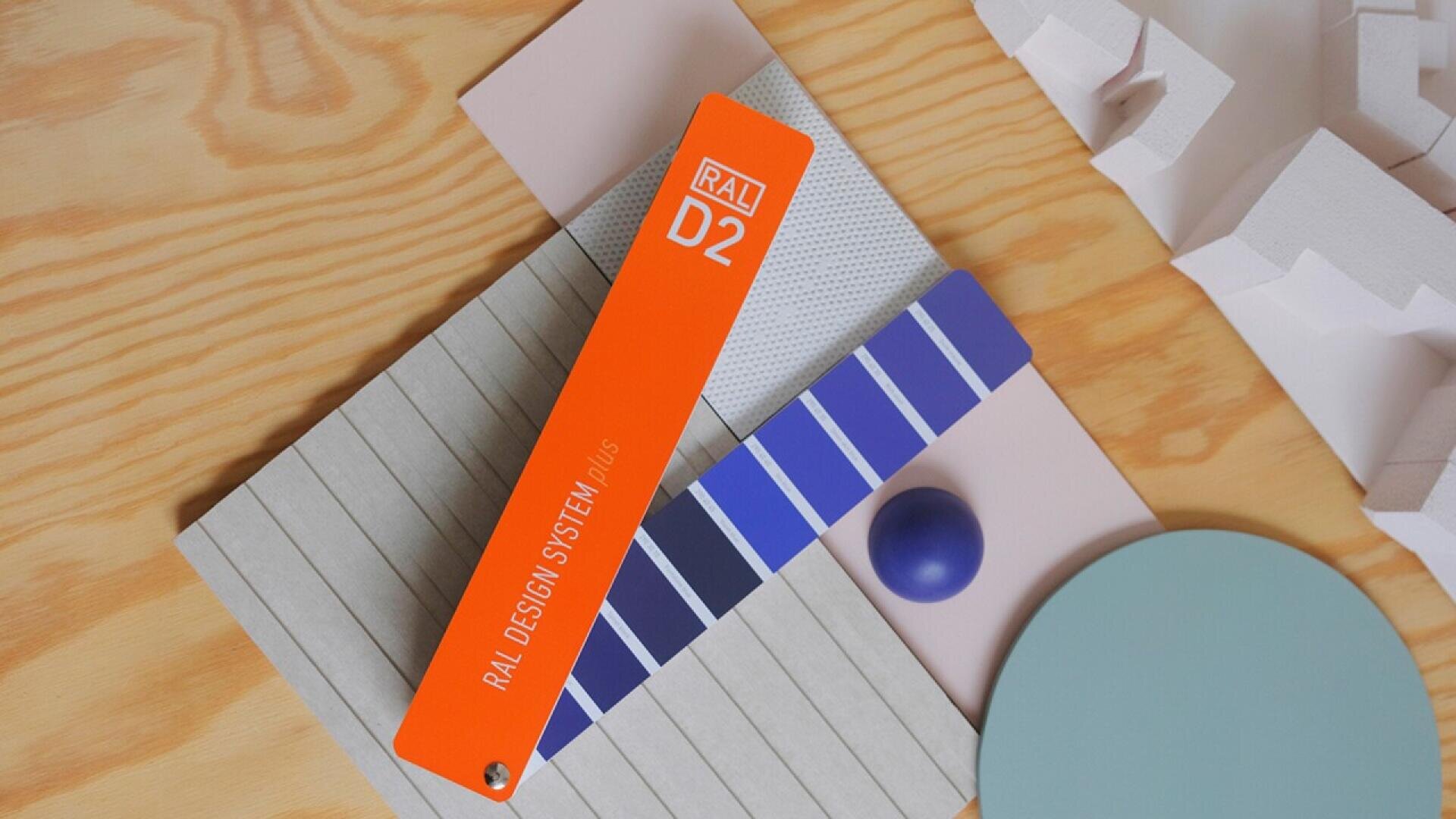 A RAL D2 color fan deck with an orange cover and blue color samples lies on a textured wooden surface, surrounded by geometric shapes in pastel colors and a purple sphere.