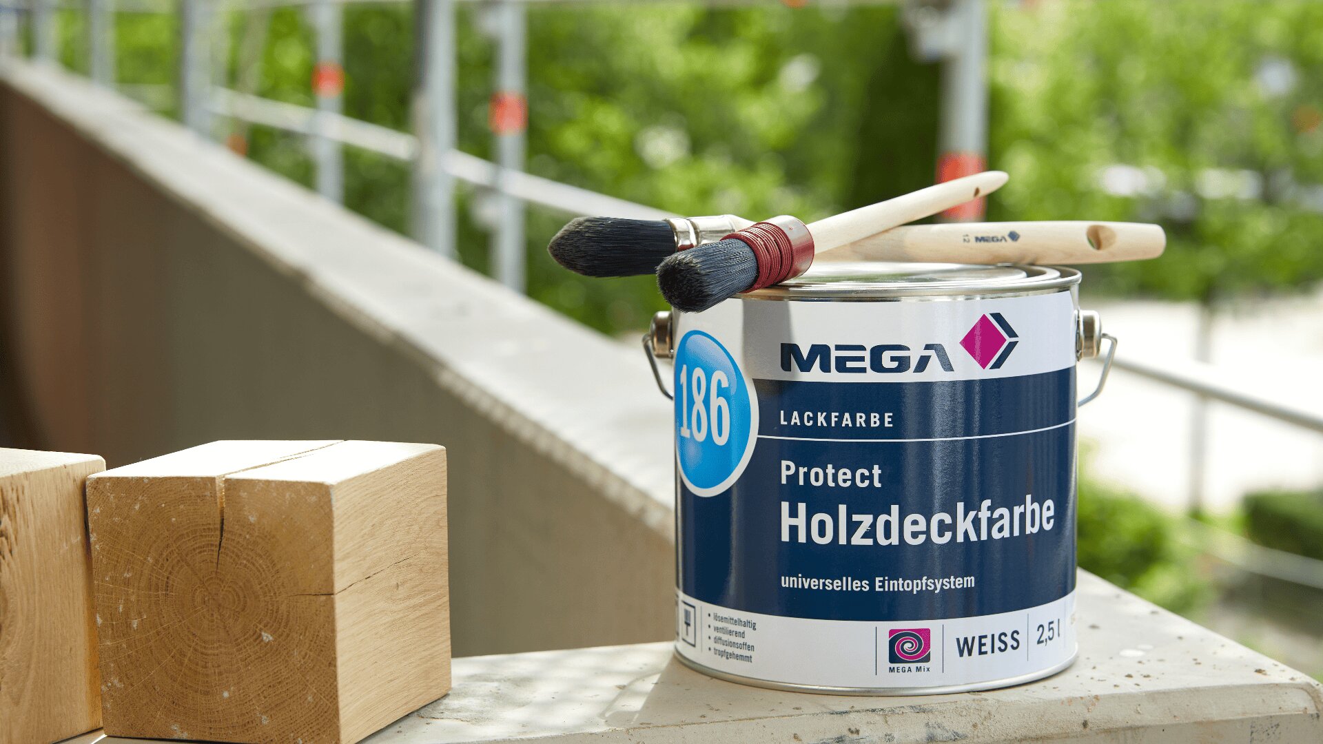 Eine Dose Holzdeckfarbe der Marke Mega steht neben zwei Pinseln und zwei Holzklötzen auf einem Balkonsims, im unscharfen Hintergrund sind grüne Bäume zu sehen.