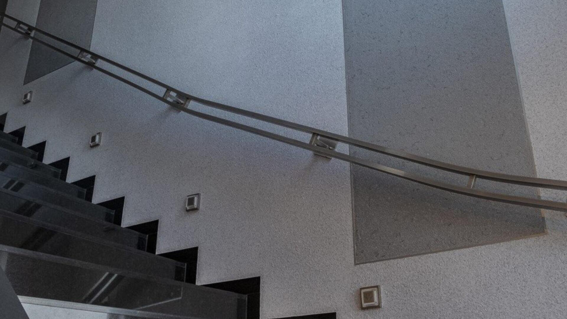 A modern interior staircase with black steps and a sleek metal handrail, set against a textured gray wall with square light fixtures.