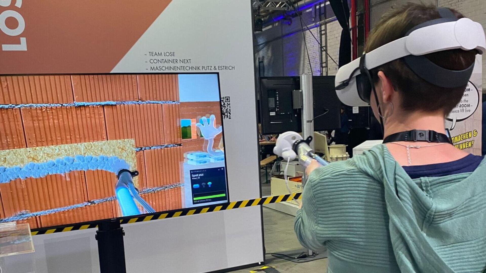 A person wearing a VR headset and holding controllers interacts with a large screen that displays a bricklaying simulation in a modern exhibition or event space.