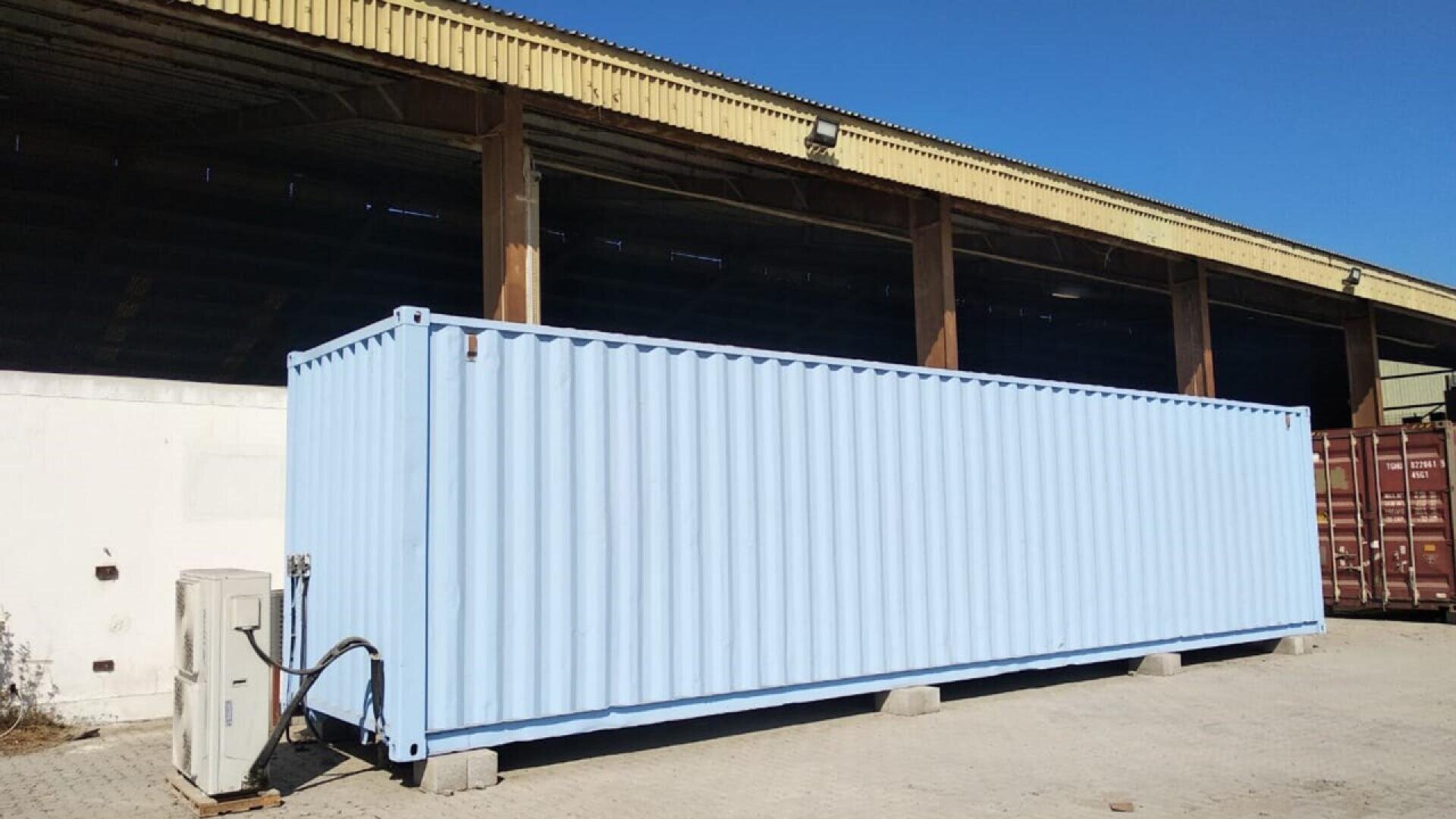 A large, light blue shipping container sits on concrete blocks outside an industrial warehouse, next to an air conditioning unit under a clear blue sky.