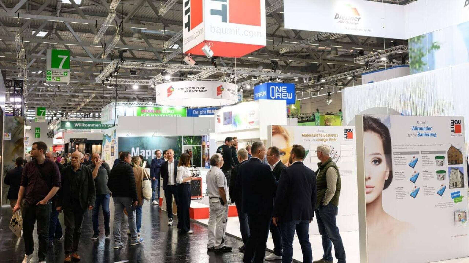 People walk through a busy exhibition hall with various booths and banners, including Baumit and other companies. Attendees are engaged in conversations and browsing displays in a well-lit, modern setting.