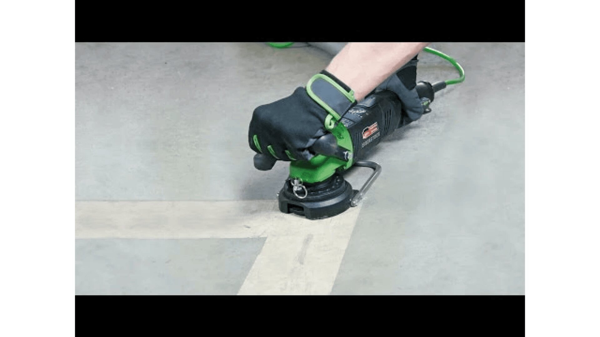 Eine Person mit schwarzen Handschuhen verwendet ein grün-schwarzes Elektrowerkzeug, um entlang der Kante einer Maßbandlinie auf einem Betonboden zu schleifen oder zu schneiden.