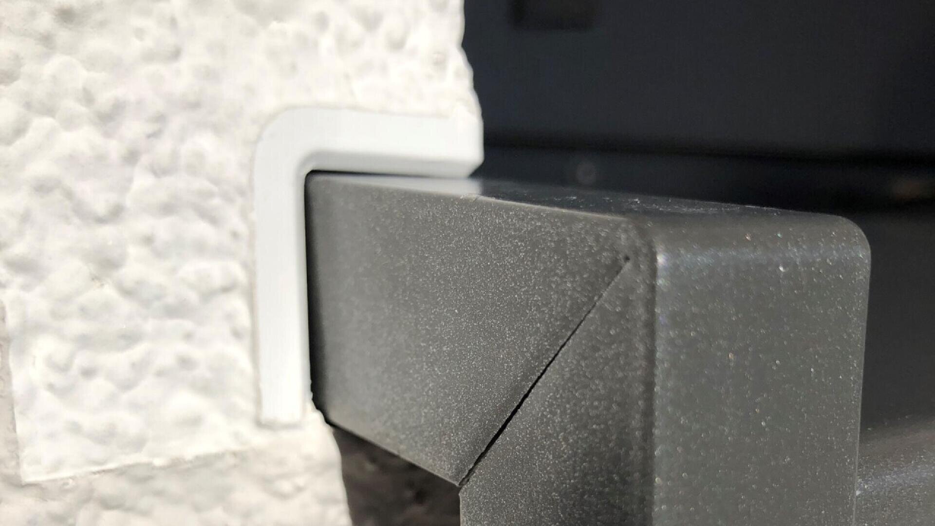 Close-up of a textured white wall corner with a white plastic edge guard, adjacent to a dark gray, smooth, rectangular surface. The surfaces meet at a right angle.
