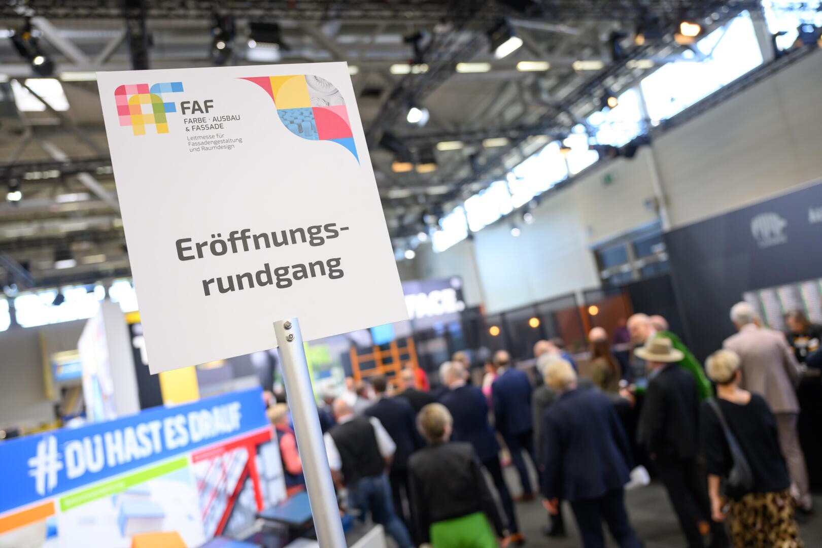 Ein Schild mit der Aufschrift „Eröffnungsrundgang“ steht bei einer Indoor-Veranstaltung im Fokus, im Hintergrund sind eine verschwommene Menschenmenge und Messestände zu sehen. Die Atmosphäre ist geschäftig und professionell.