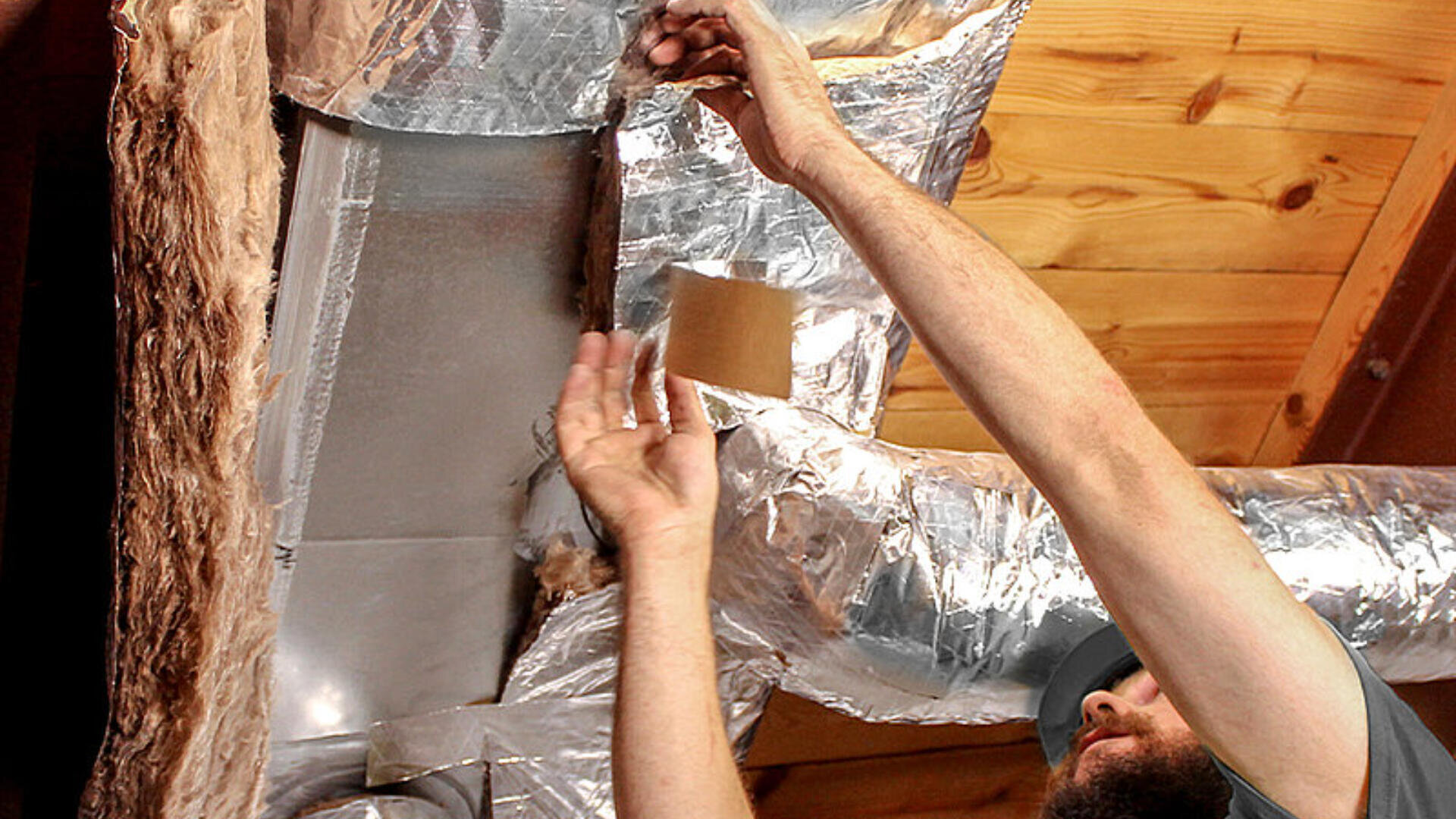 Eine Person bringt Metallband an, um die Isolierung rund um die Rohrleitungen an einer Decke mit Holzverkleidung abzudichten.